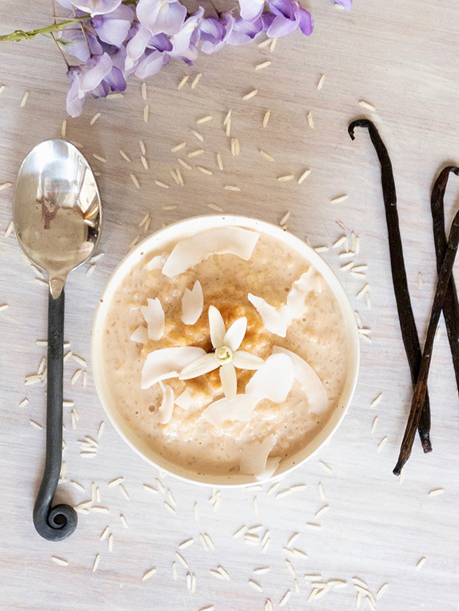 White bowl of creamy rice pudding with cinnamon and shredded coconut on top. Black spoon and vanilla beans on the side.